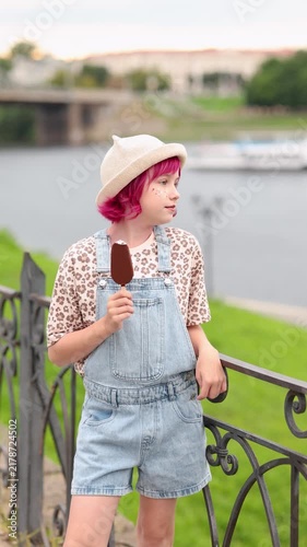 Smiling child with pink hair and straw hat holds chocolate coated ice cream on stick while standing by ornate metal railing beside calm river, girl wears leopard print shirt under light denim