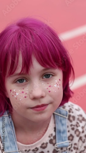 Bright portrait of smiling pregnant woman with pink hair, beige knit hat, heart stickers, freckles, and denim overall strap, posed against soft pink backdrop with warm gentle light