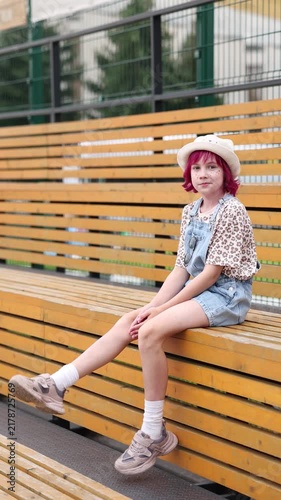 Child with pink hair sits alone on outdoor wooden bleachers wearing light hat, patterned short sleeve shirt, and denim overalls, relaxed neutral expression, casual sneakers and white socks visible