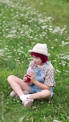 Young child with bright pink hair and straw hat sits cross legged in grassy wildflower meadow, smiling at camera while holding clear cup of red beverage with straw, wearing denim overalls and