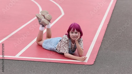 Young girl with bright pink hair and face glitter lies on stomach on pink outdoor sports court, propping head on hand and smiling toward camera, wearing denim overalls, leopard print shirt, white