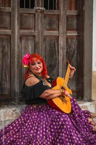 A full-figured Latina Gypsy woman playing traditional Gypsy songs on the guitar; an old yellow house is visible in the background.
