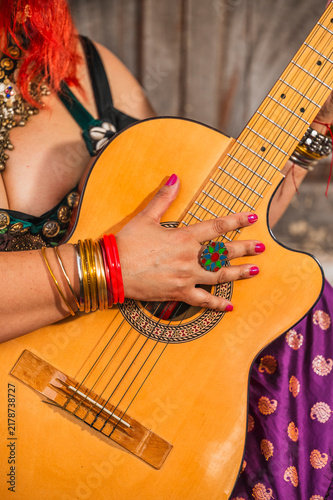 A full-figured Latina Gypsy woman playing traditional Gypsy songs on the guitar; an old yellow house is visible in the background.
