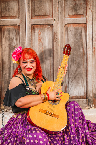 A full-figured Latina Gypsy woman playing traditional Gypsy songs on the guitar; an old yellow house is visible in the background.

