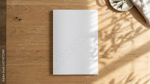 Blank book cover mockup on a wooden table with soft sunlight and leaf shadows, top down flat lay view of white magazine template with minimalist ceramic dish and linen cloth