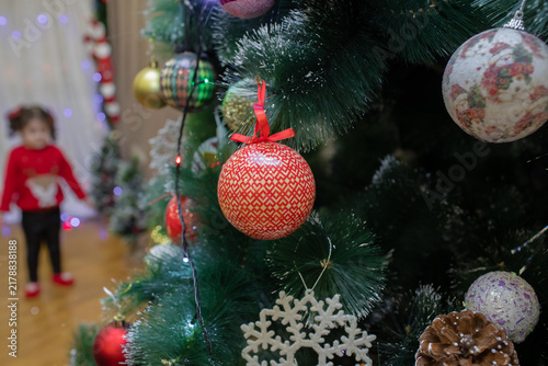 In the blurred background, a toddler girl stands near lights in cozy home settings Beautiful red ornament hanging on a lush green Christmas tree decorated with colorful festive baubles and snowflakes.