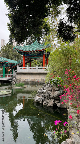 traditional chinese pavilion reflecting serene garden pond from the side with flowers and trees in green and red tones, landscape photography travel architecture.
