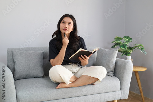Obraz na płótnie A young woman is focused on writing in a notebook while sitting on a comfortable sofa; she appears to be deep in thought and reminiscing, radiating creativity and inspiration as she looks upward