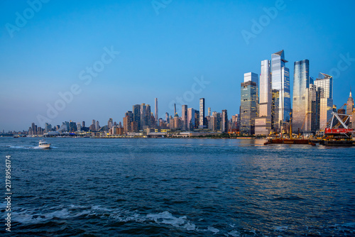 Midtown Manhattan skyline at twilight viewed over water in New York. Highrise modern towers illuminate evening sky above blue river water with small boat passing by.