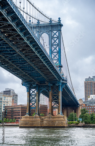 Blue steel structure of Manhattan Bridge viewed from water level in New York. Low angle perspective of massive bridge support column and truss framework above river water under overcast sky.