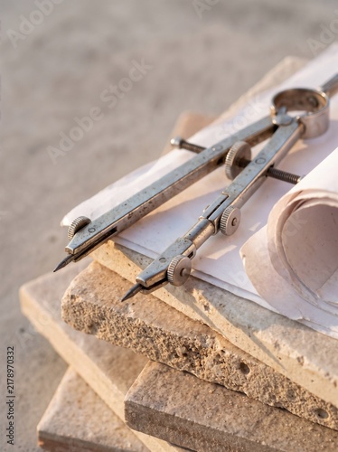 Classic metal drafting compass on stacked stone tiles with a rolled paper blueprint, precise geometric planning and traditional architectural design, warm sunlight outdoor background
