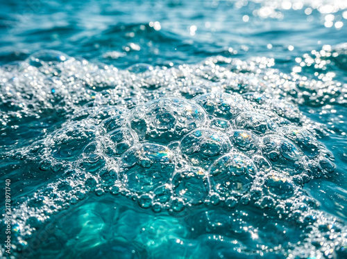 Blue ocean water surface with white foam bubbles and sparkling sunlight reflections.