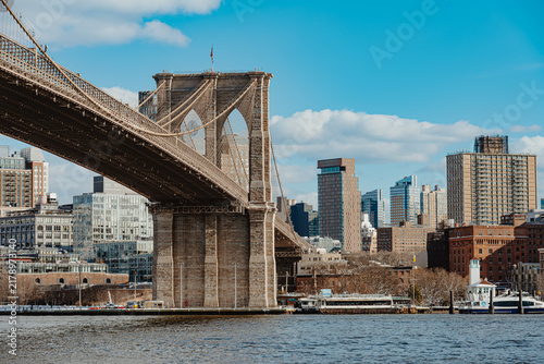 Brooklyn Bridge tower standing over East River, New York. Wide view of massive stone pier and suspension cables against blue sky and city buildings.