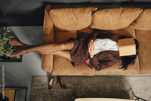 A woman lounges comfortably on a stylish couch, dressed in a suit. She places a book over her face, enjoying a moment of quiet in a tastefully decorated living space during the day