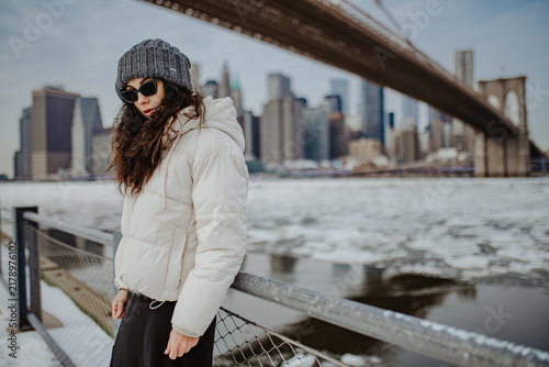 Three quarter view of woman standing by riverfront railing, New York. Focused pose wearing dark eyewear, winter coat, grey cap against icy water and city buildings.