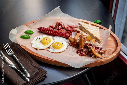 Breakfast plate with eggs, sausage, and bread on a table. A breakfast meal includes eggs, sausage, bacon, and bread on a wooden plate at a cafe.