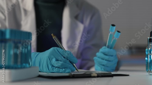 Scientist in a lab coat and blue gloves writing notes on a clipboard and holding test tubes with blue liquid, researching a new coronavirus vaccine or developing a drug