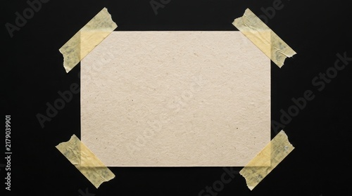 A textured beige piece of paper secured to a solid black background with four strips of yellow masking tape at each corner