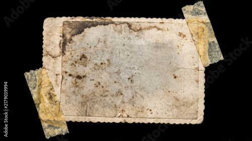 An aged and weathered vintage paper frame with scalloped edges held by pieces of discolored adhesive tape against a black background