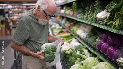 An elderly man is selecting fresh broccoli in a grocery store. He examines the produce while standing near the vegetable section