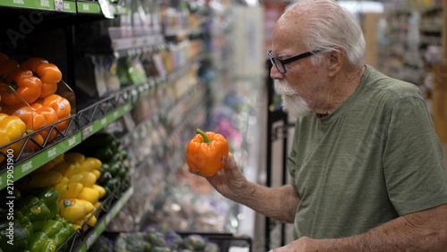 Senior man is shopping in a grocery store and looking at colorful bell peppers in the produce section while making choices