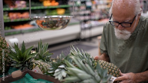 An elderly man inspects fresh pineapples while shopping in a grocery store during the daytime. He focuses on quality and freshness
