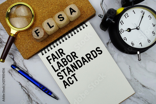 FLSA Fair Labor Standards Act text written on notepad with magnifying glass and alarm clock on marble background
