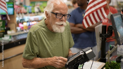 An elderly man shops at a grocery store. He stands at the checkout, paying for his items. Other customers are nearby