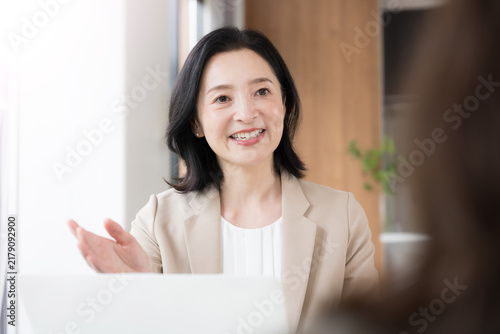Naklejka 笑顔で顧客と対面相談する50代女性、オフィスで説明や提案を行うビジネスウーマン コミュニケーションや商談などのビジネスイメージ