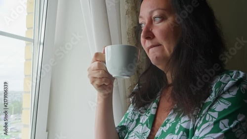 Woman in green floral shirt holds white mug while looking out window, capturing peaceful morning routine, relaxation, contemplation by natural light indoors.