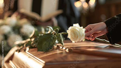 Fresh white rose being put by woman in mourning attire on top of closed coffin lid against priest with open Bible carrying out funeral service