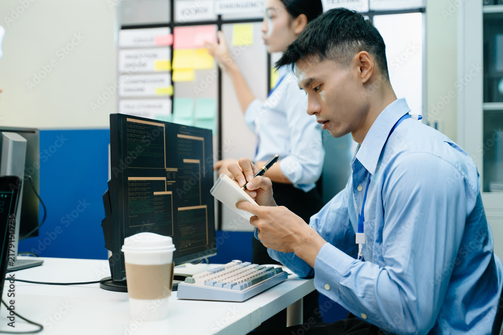 Fototapeta premium Dedicated young software engineer writing notes in a notebook while coding and monitoring data systems