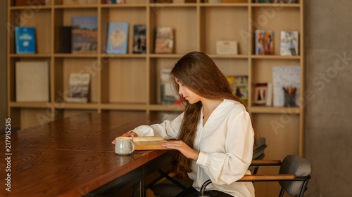 Fotografie Young woman engrossed in a book while enjoying a warm beverage at a wooden table
