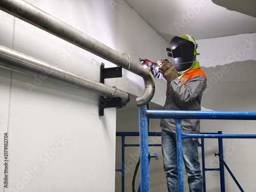 A wide-angle, low-angle shot of a technician standing on mobile scaffolding to perform pipe welding near the ceiling in a spacious cleanroom facility with a green epoxy floor 