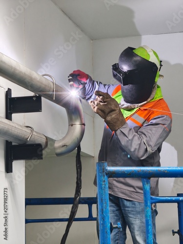 A low-angle shot of an industrial welder working on scaffolding, using TIG welding on a stainless steel pipe joint, creating a bright welding arc light flare while wearing a flip-up welding mask