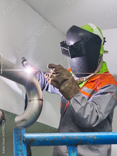 A low-angle shot of an industrial welder working on scaffolding, using TIG welding on a stainless steel pipe joint, creating a bright welding arc light flare while wearing a flip-up welding mask