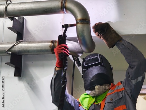 A low-angle shot of a professional welder wearing a welding helmet and protective gloves performing TIG welding on an overhead stainless steel pipe joint in an industrial facility.