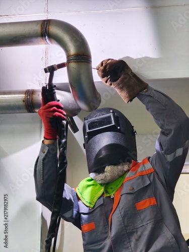 A low-angle shot of a professional welder wearing a welding helmet and protective gloves performing TIG welding on an overhead stainless steel pipe joint in an industrial facility.
