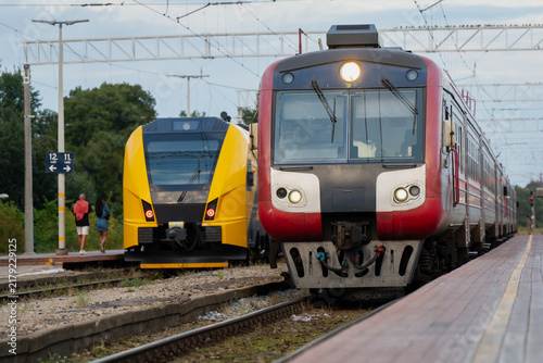 Foto Headlights glow against overcast station light as an older red train arrives beside a waiting yellow regional unit, two generations of fleet at one platform