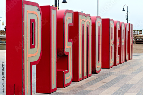 Картината върху платно Large red Pismo Beach letter sign stands on an oceanfront plaza with palm trees and decorative street lamps beneath an overcast sky
