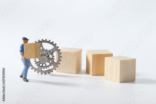 Miniature worker carrying a box beside gears and wooden blocks