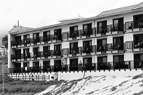 Photography Multi-story oceanfront building with rows of balconies, black railings and people overlooking sandy dunes in Pismo Beach, California