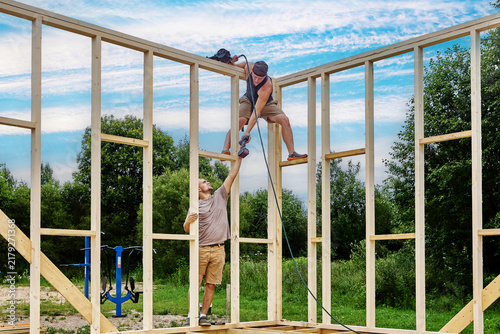 Foto Teamwork during house framing as young worker passes cordless drill to colleague standing on wooden wall frame