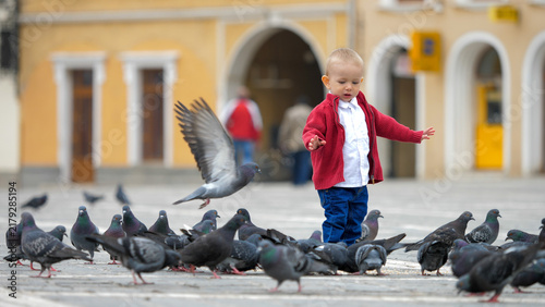 Fotografía Little blond baby child walking between pigeons that peck on city street