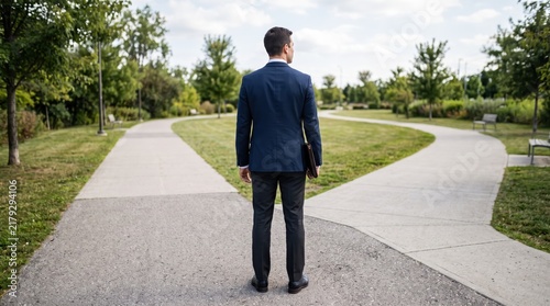 Businessman standing at fork in road facing decision making choice, man in suit holding briefcase looking at diverging path in park, career direction crossroads metaphor