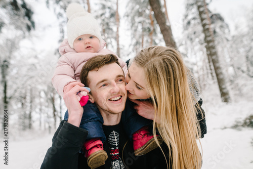 Konstfotografi Happy parents in snowy forest, mother tenderly kissing smiling father who carries their little daughter on his shoulders
