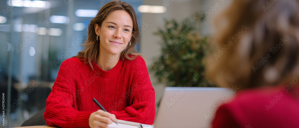 Fototapeta premium Young woman in red sweater attentively listening and taking notes during a casual meeting with colleague