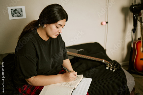 Foto Young biracial woman writing song lyrics in notebook near acoustic guitar on bed
