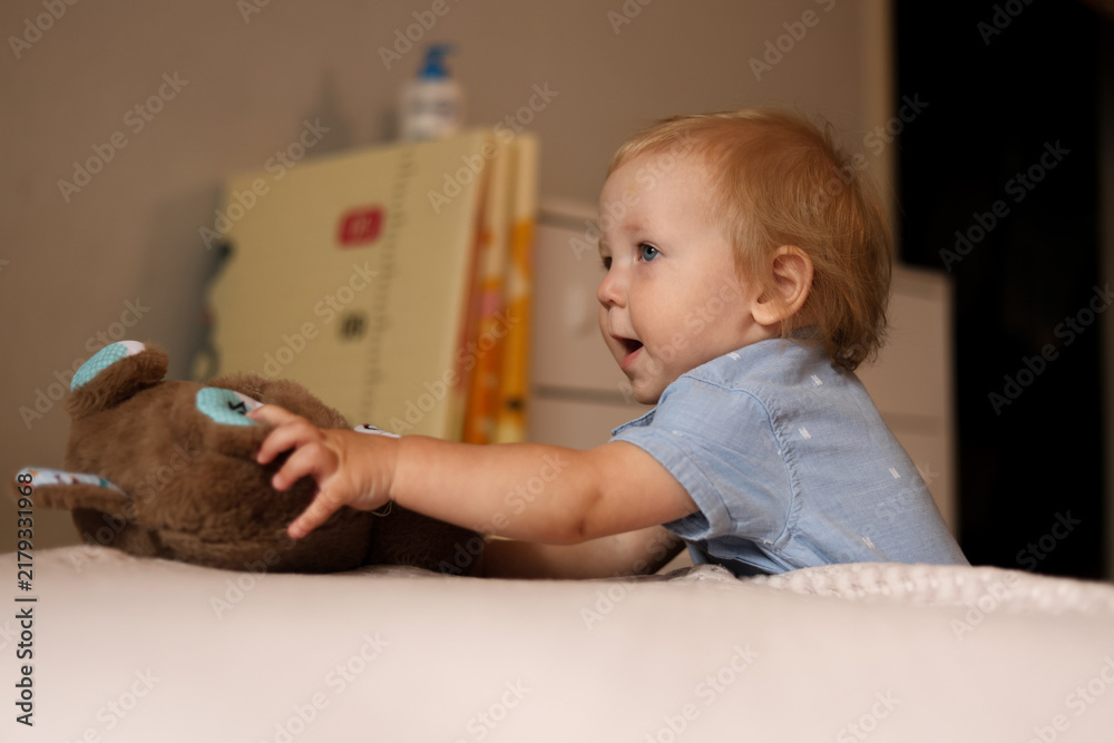 Naklejka premium Curious toddler reaching for a soft teddy bear on a bed