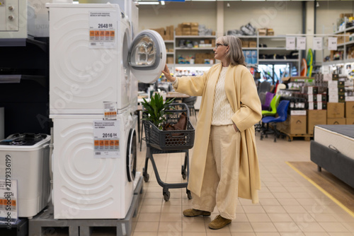 Senior woman inspecting a washing machine, comparing appliances in a modern department store